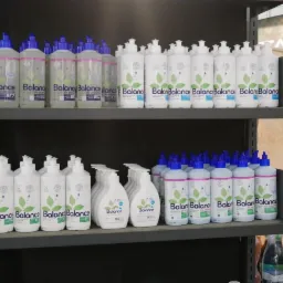 Rzędy białych butelek z niebieskimi i białymi nakrętkami, z etykietami 'Balance' z zielonym motywem liści, ustawione na ciemnych półkach regału.