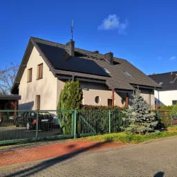 Dom jednorodzinny z panelami słonecznymi na dachu, widoczny z ulicy przez zielone ogrodzenie; na podjeździe zaparkowany czarny samochód.