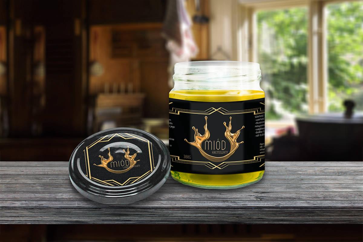 Słoik kremowego miodu z czarną etykietą z motywem złotych kropli i pokrywka ze zbliżonym wzorem, na drewnianym blacie z rozmytym tłem wnętrza.