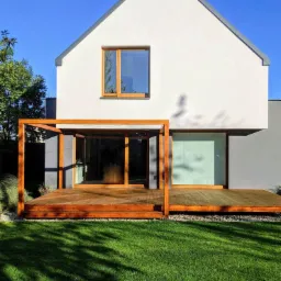 Nowoczesny dom z białym tynkiem i drewnianą pergolą nad tarasem, widok z ogrodu z intensywnie zielonym trawnikiem i cieniami drzew.