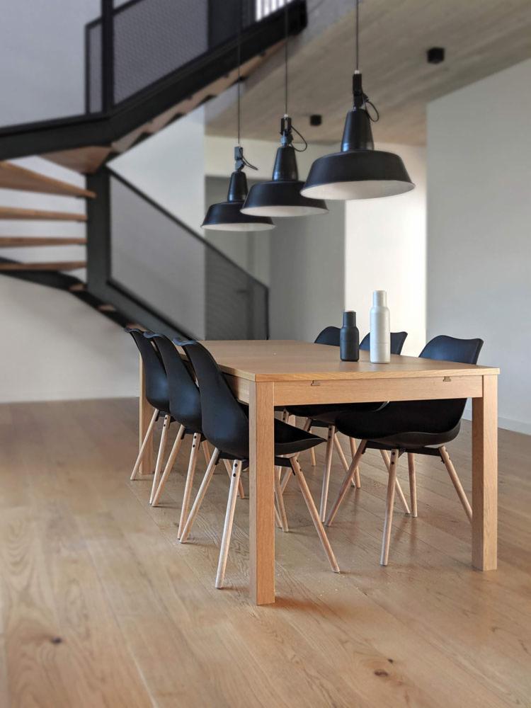 Jadalnia z drewnianym stołem, czarnymi krzesłami i trzema wiszącymi lampami w stylu industrialnym, schody z metalową balustradą w tle.