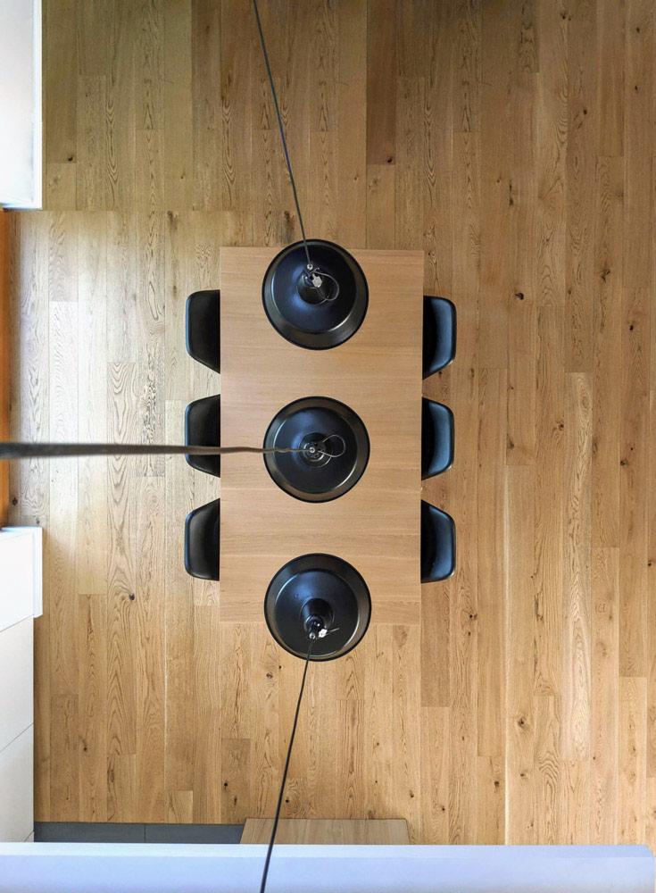 Widok z góry na stół z jasnego drewna z trzema czarnymi lampami wiszącymi, otoczony czarnymi krzesłami na tle drewnianej podłogi.