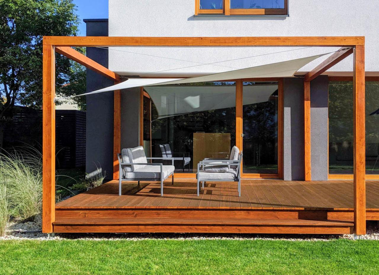 Drewniany taras z pergolą i żaglem przeciwsłonecznym, dwie szare sofy z poduszkami, trawnik i biały żwir.