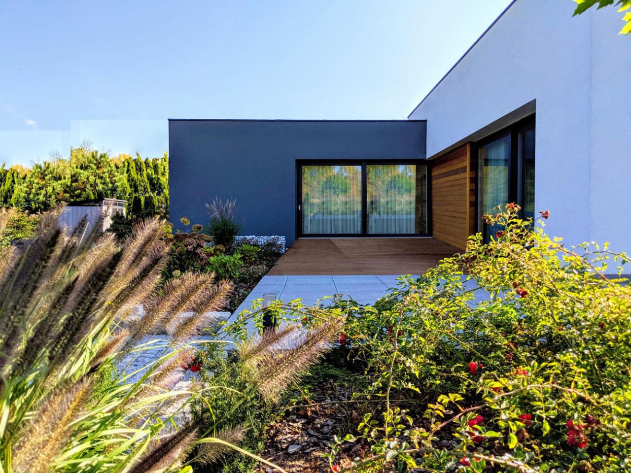 Nowoczesny dom jednorodzinny z płaskim dachem, antracytową ścianą i drewnianym tarasem, otoczony zielenią i ozdobnymi trawami w słoneczny dzień.