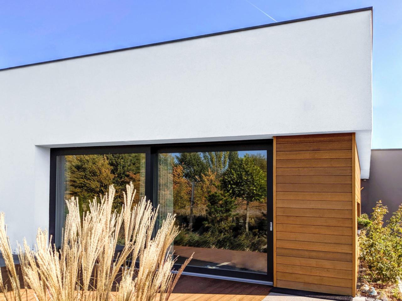 Nowoczesny dom z białym tynkiem, dużym przesuwnym oknem z czarną ramą i drewnianą elewacją, z widocznymi elementami krajobrazu w odbiciu okna.