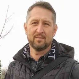 Portret mężczyzny z brodą w kurtce z kapturem, z torbą przewieszoną przez ramię, na tle rozmytego krajobrazu.