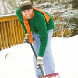 Uśmiechnięty mężczyzna w czapce i szaliku odśnieża śnieg łopatą na tle zasypanych śniegiem drzew i drewnianego płotu.