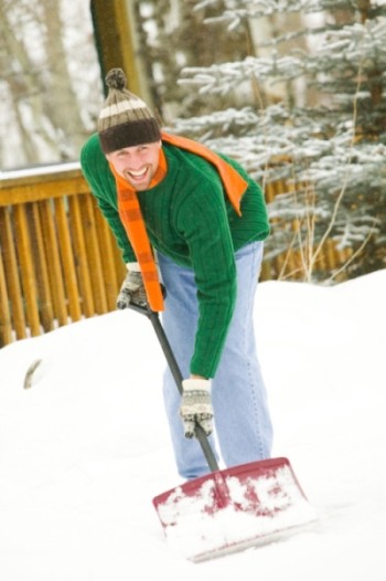 Uśmiechnięty mężczyzna w czapce i szaliku odśnieża śnieg łopatą na tle zasypanych śniegiem drzew i drewnianego płotu.