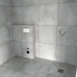 Wykończona łazienka z szarymi, kwadratowymi płytkami na ścianach i podłodze, widoczne otwory na armaturę i odpływ podtynkowy.