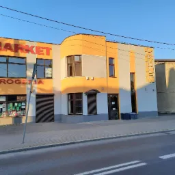 Dwukondygnacyjny budynek z napisem 'MARKET DROGERIA' w kolorach pomarańczowo-szarych, z widoczną fasadą po remoncie, obejmującą nowe okna i drzwi wejściowe.