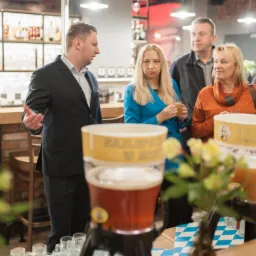 Grupa osób w barze podczas imprezy, z dystrybutorami piwa i szklankami na pierwszym planie, mężczyzna w garniturze gestykuluje.