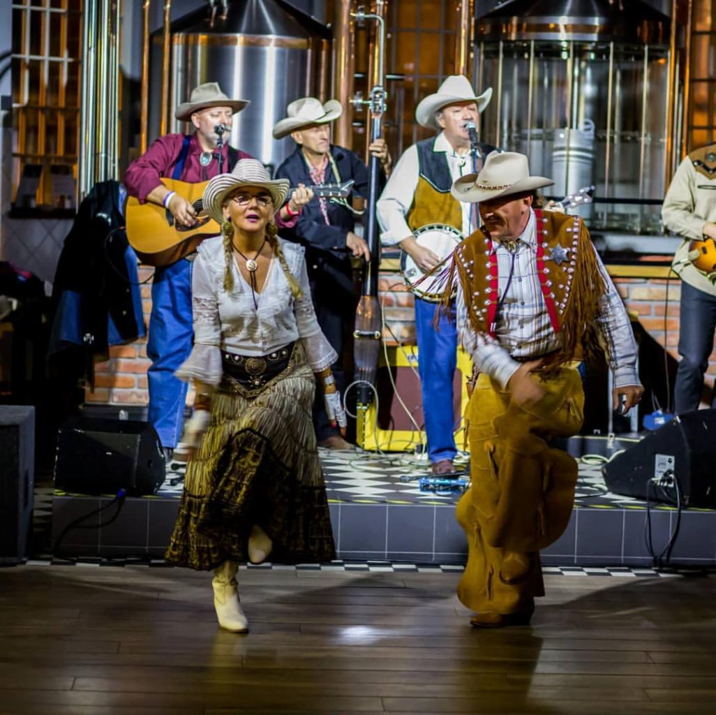Zespół muzyczny w strojach kowbojskich występuje na scenie z instrumentami, w tle stalowe kadzie browarnicze i ceglana ściana