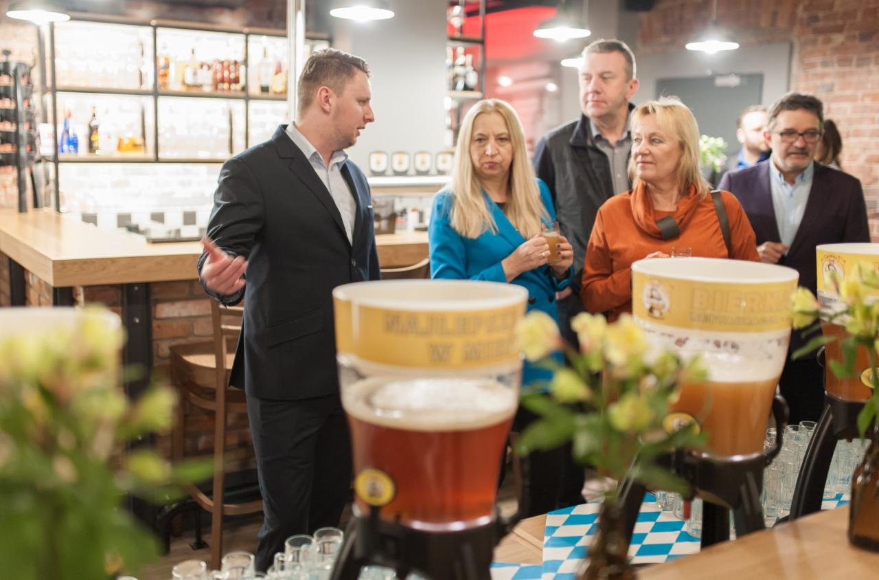 Grupa osób w barze podczas imprezy, z dystrybutorami piwa i szklankami na pierwszym planie, mężczyzna w garniturze gestykuluje.