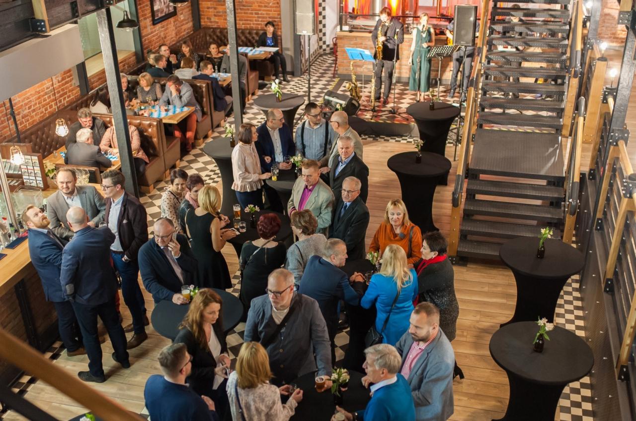 Wnętrze loftu z ceglanymi ścianami, gdzie odbywa się wydarzenie firmowe: goście stoją przy czarnych stolikach koktajlowych z kwiatami, w tle gra zespół muzyczny, a na podłodze widać wzór szachownicy.