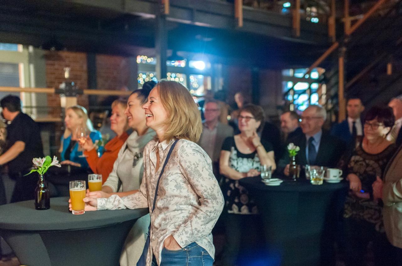 Uczestnicy wydarzenia w industrialnym wnętrzu, kobieta w kwiecistej koszuli trzyma piwo, uśmiechnięta, w tle grupa osób przy stolikach.