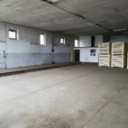 Wnętrze magazynu z betonową podłogą i ścianami, małe okna z kratami, rzędy drewnianych palet w prawym rogu.