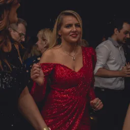Kobieta w czerwonej, cekinowej sukience tańczy na imprezie, w tle widoczni inni goście.