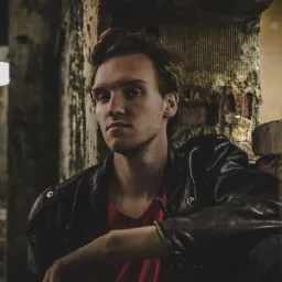 Portret młodego mężczyzny w skórzanej kurtce na tle surowej ściany z cegieł i betonowych elementów, oświetlony lampą z boku.