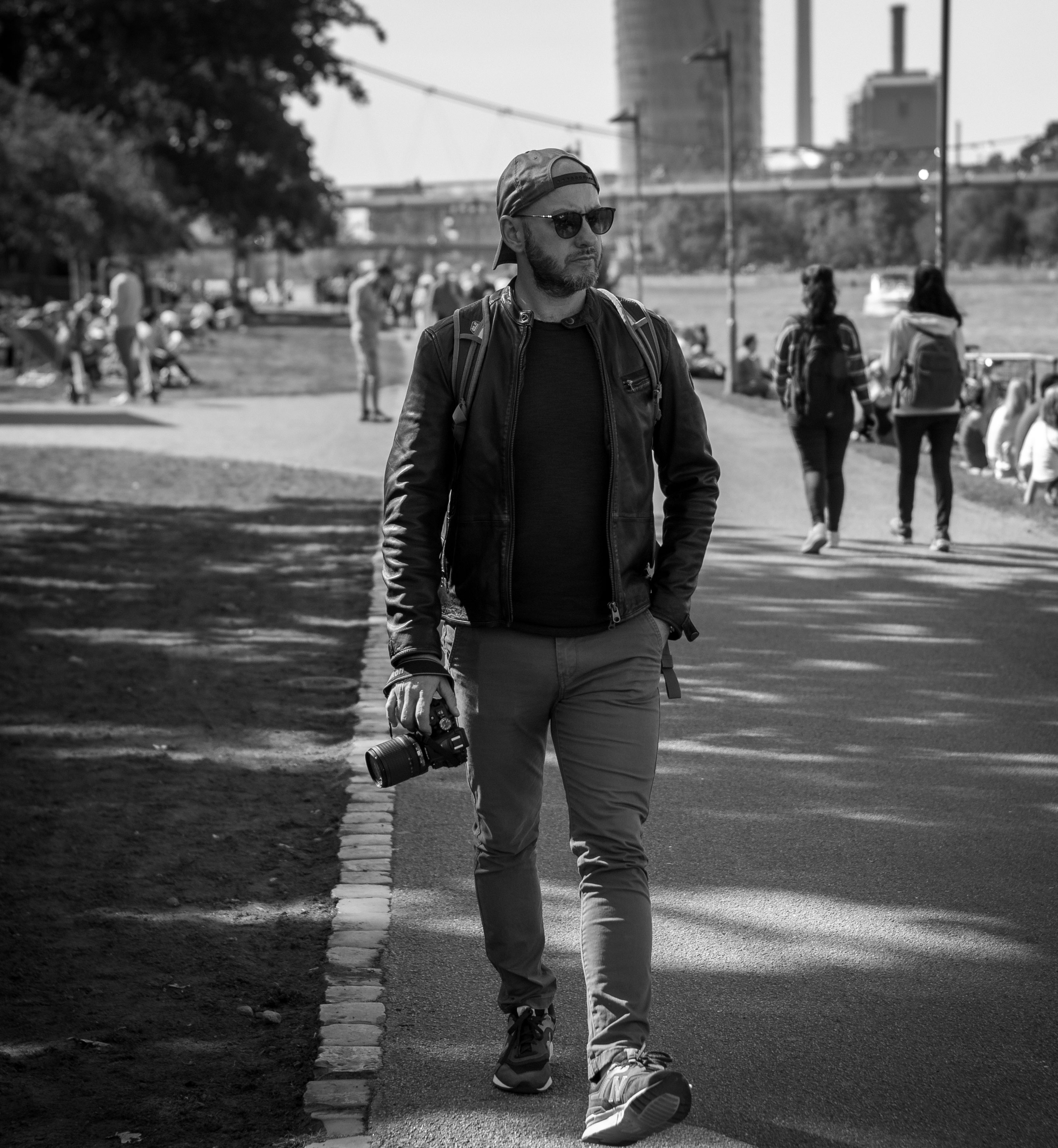 Czarno-białe zdjęcie fotografa z aparatem spacerującego po parku w okularach przeciwsłonecznych i czapce z daszkiem, w tle widoczni ludzie i miejski krajobraz z kominami.