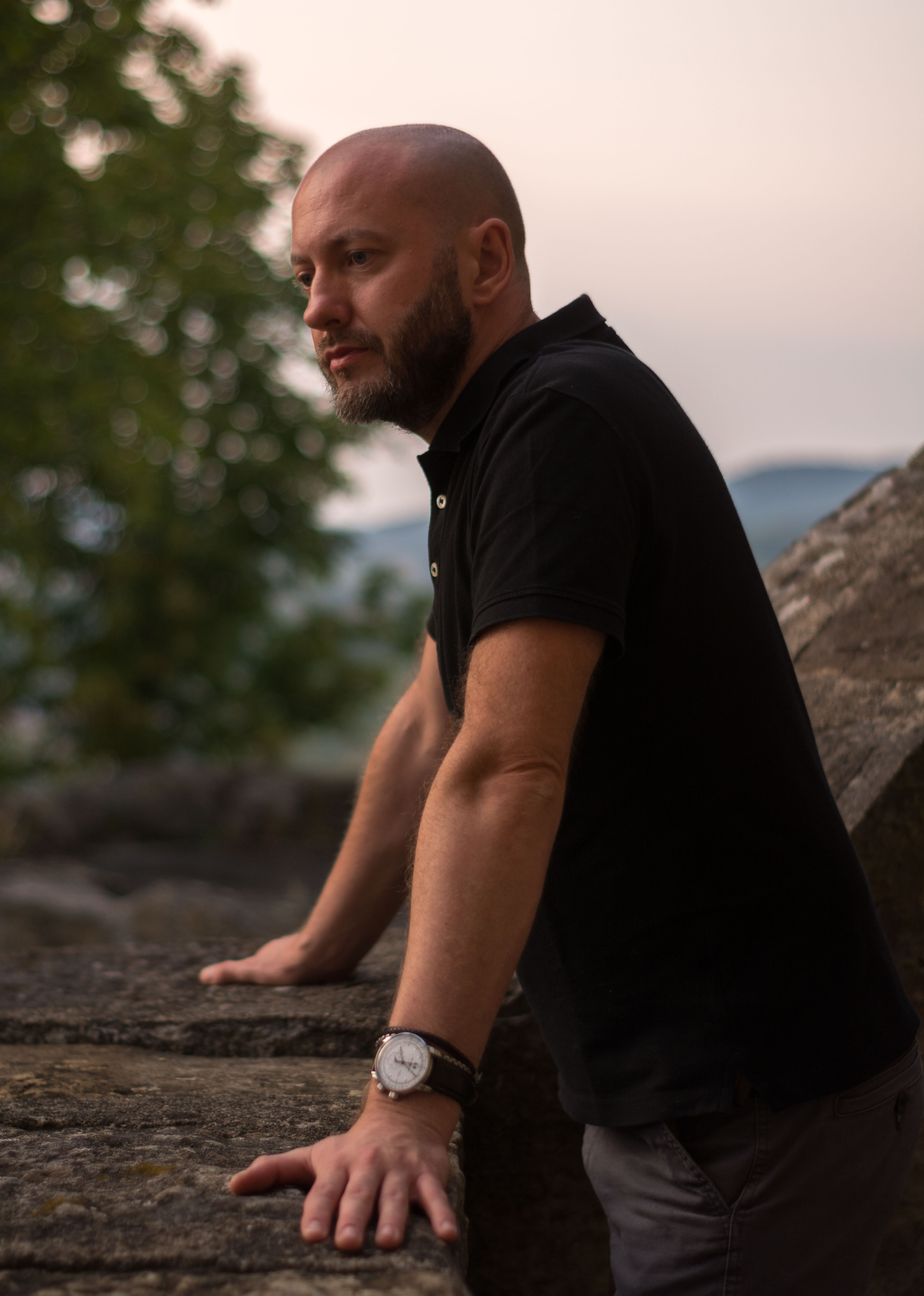 Mężczyzna z brodą w czarnej koszulce polo, oparty o kamienny mur z widocznym zegarkiem na nadgarstku, zamyślony, w tle rozmyte drzewa i wzgórza.