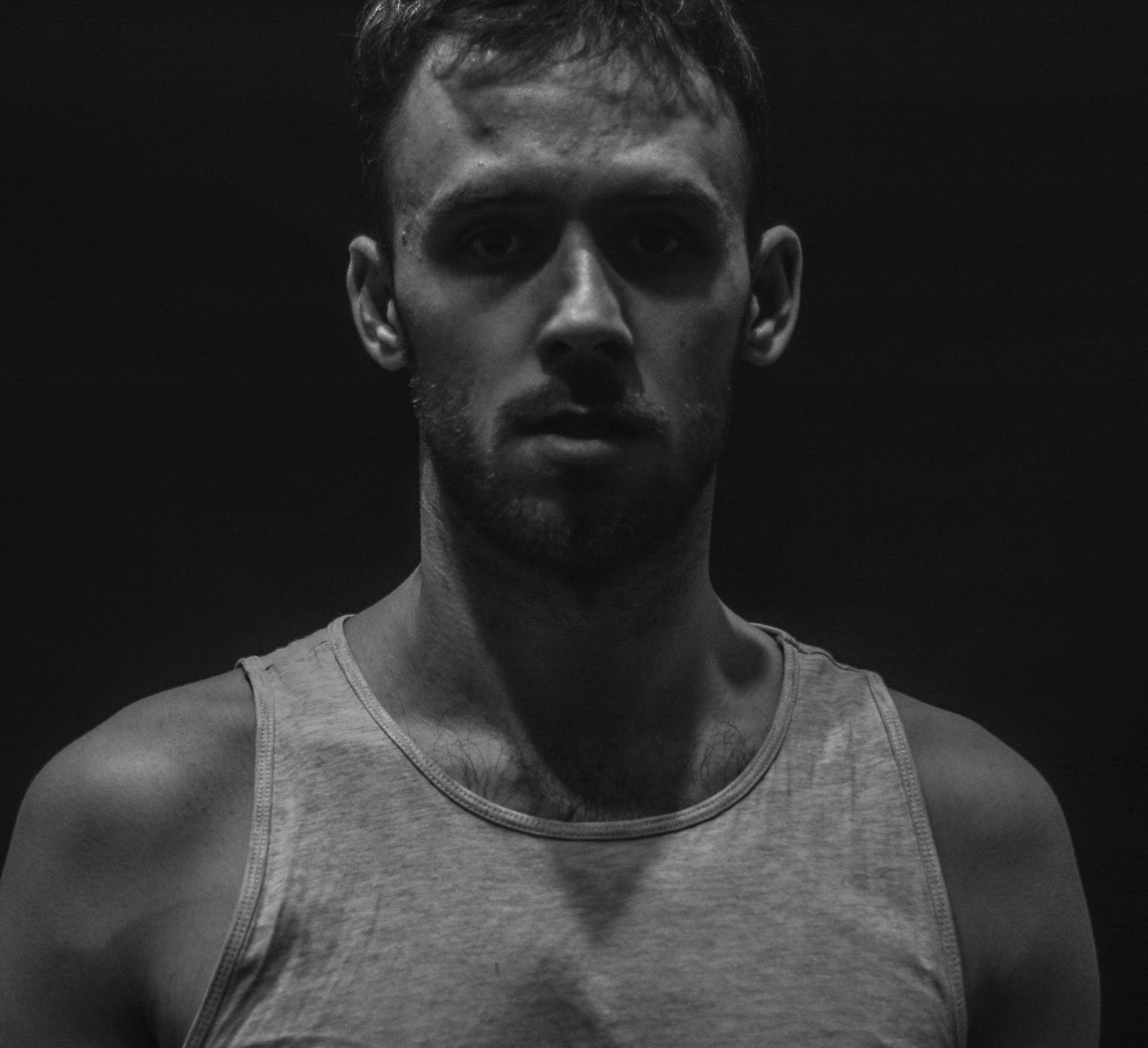 Czarno-biały portret młodego mężczyzny z krótką brodą, oświetlonego od góry, na ciemnym tle.