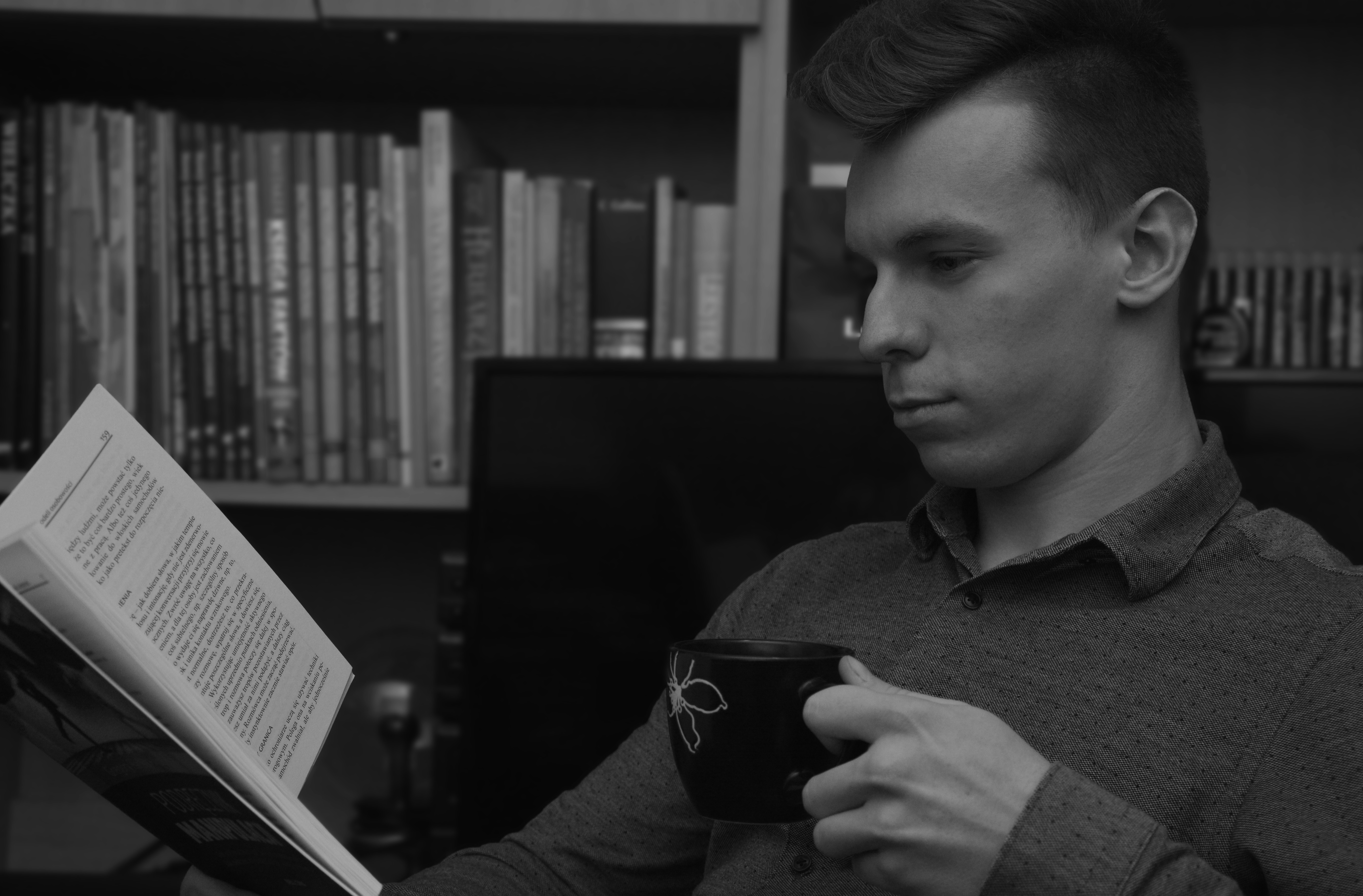 Mężczyzna w koszuli czyta książkę i trzyma kubek z kwiatowym wzorem na tle półki z książkami, zdjęcie czarno-białe.