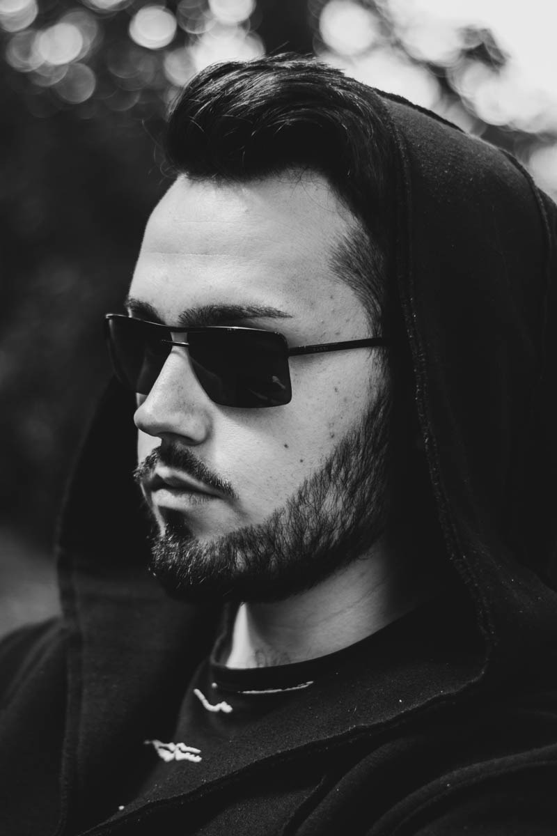 Portret en face młodego mężczyzny z brodą w kapturze i okularach przeciwsłonecznych, czarno-biały, z rozmytym tłem bokeh.