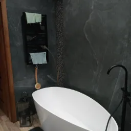 Nowoczesna łazienka z wolnostojącą białą wanną na tle ściany wykończonej grafitową płytką imitującą kamień, czarna armatura i drabinka na ręczniki.