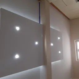 Białe panele ścienne z oświetleniem punktowym LED, oddzielone pionowym elementem architektonicznym, widok na fragment sufitu i okna.