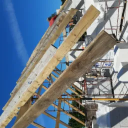 Wznoszenie konstrukcji dachu z drewnianych belek na budynku z białych bloczk&oacute;w, widok z dołu na błękitne niebo z smugą kondensacyjną.