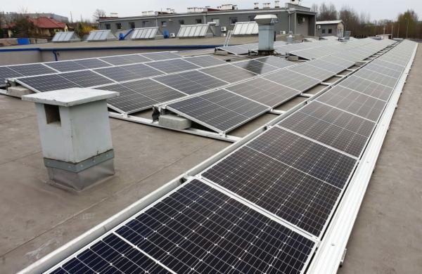 Rząd paneli słonecznych na płaskim dachu budynku przemysłowego, widoczny szary komin wentylacyjny i dalsze rzędy paneli.
