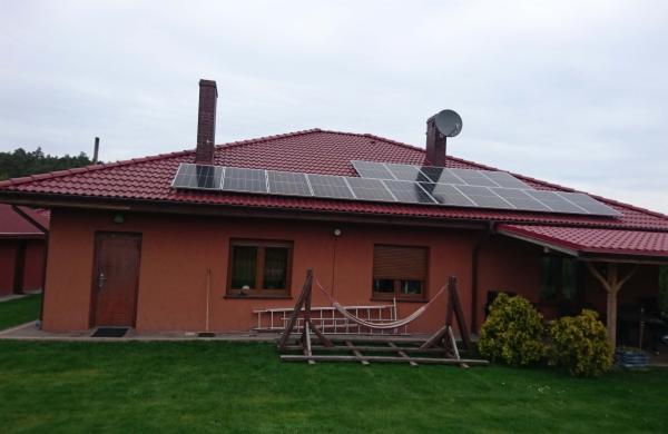 Dom jednorodzinny z panelami słonecznymi na czerwonym dachu, widoczny komin, antena satelitarna, drabina oparta o ścianę i hamak w ogrodzie.