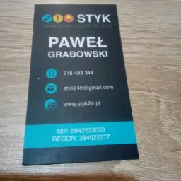 Wizytówka firmy elektrycznej 'STYK' z danymi kontaktowymi Pawła Grabowskiego, obejmująca instalacje elektryczne i remonty, leżąca na drewnianym blacie.