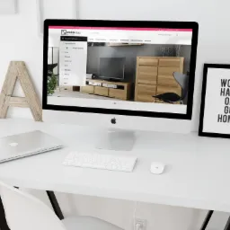 Ekran komputera iMac prezentuje stronę internetową sklepu meblowego 'mebledąb' z kolekcją mebli dębowych. Na biurku obok laptop, klawiatura, mysz oraz drewniana litera 'A' i plakat z napisem 'WORK...