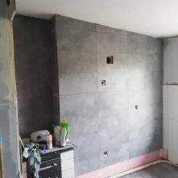 Ściana wyłożona dużymi, szarymi płytkami ceramicznymi z widocznymi otworami na gniazdka elektryczne i puszkę. Na pierwszym planie widoczny fragment pieca kaflowego z workami zaprawy.