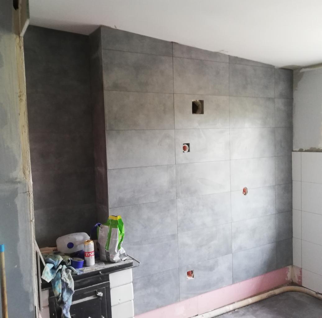 Ściana wyłożona dużymi, szarymi płytkami ceramicznymi z widocznymi otworami na gniazdka elektryczne i puszkę. Na pierwszym planie widoczny fragment pieca kaflowego z workami zaprawy.