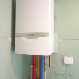 Biały piec gazowy Vaillant zamontowany na surowej ścianie z widocznymi rurami doprowadzającymi wodę w kolorach czerwonym, żółtym i niebieskim, oraz termostat na ścianie.