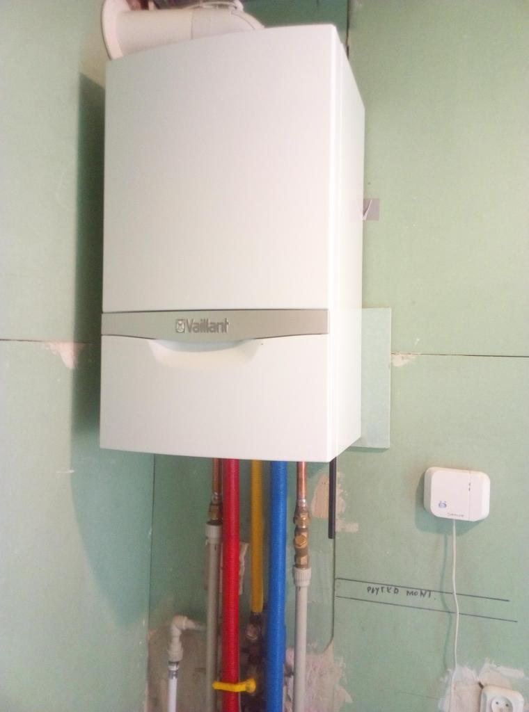 Biały piec gazowy Vaillant zamontowany na surowej ścianie z widocznymi rurami doprowadzającymi wodę w kolorach czerwonym, żółtym i niebieskim, oraz termostat na ścianie.