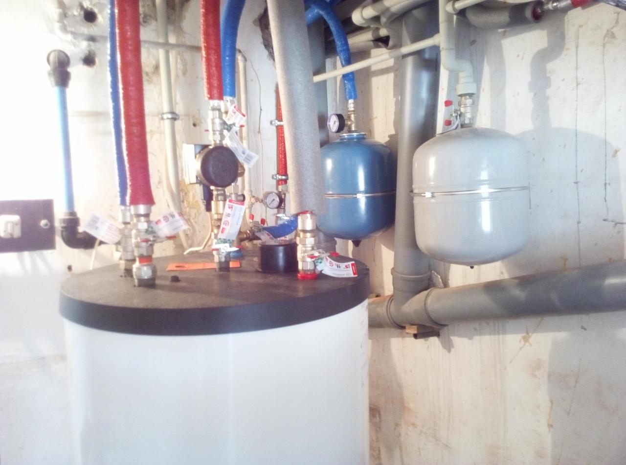 Biały zasobnik ciepłej wody użytkowej z podłączonymi rurami w kolorach czerwonym i niebieskim, widoczne także dwa zbiorniki wyrównawcze, instalacja w piwnicy o pomalowanych na biało ścianach.