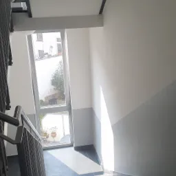Klatka schodowa z szarymi płytkami na podłodze i dolną częścią ściany, górna część ściany biała, okno na końcu biegu schodów wpuszcza światło.