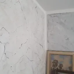 Wykończony narożnik pokoju z dwoma różnymi teksturami ścian: ściana po lewej stronie z jasną gładzią imitującą marmur z ciemnymi żyłkami, ściana po prawej stronie z szarą, rozmytą teksturą, a...