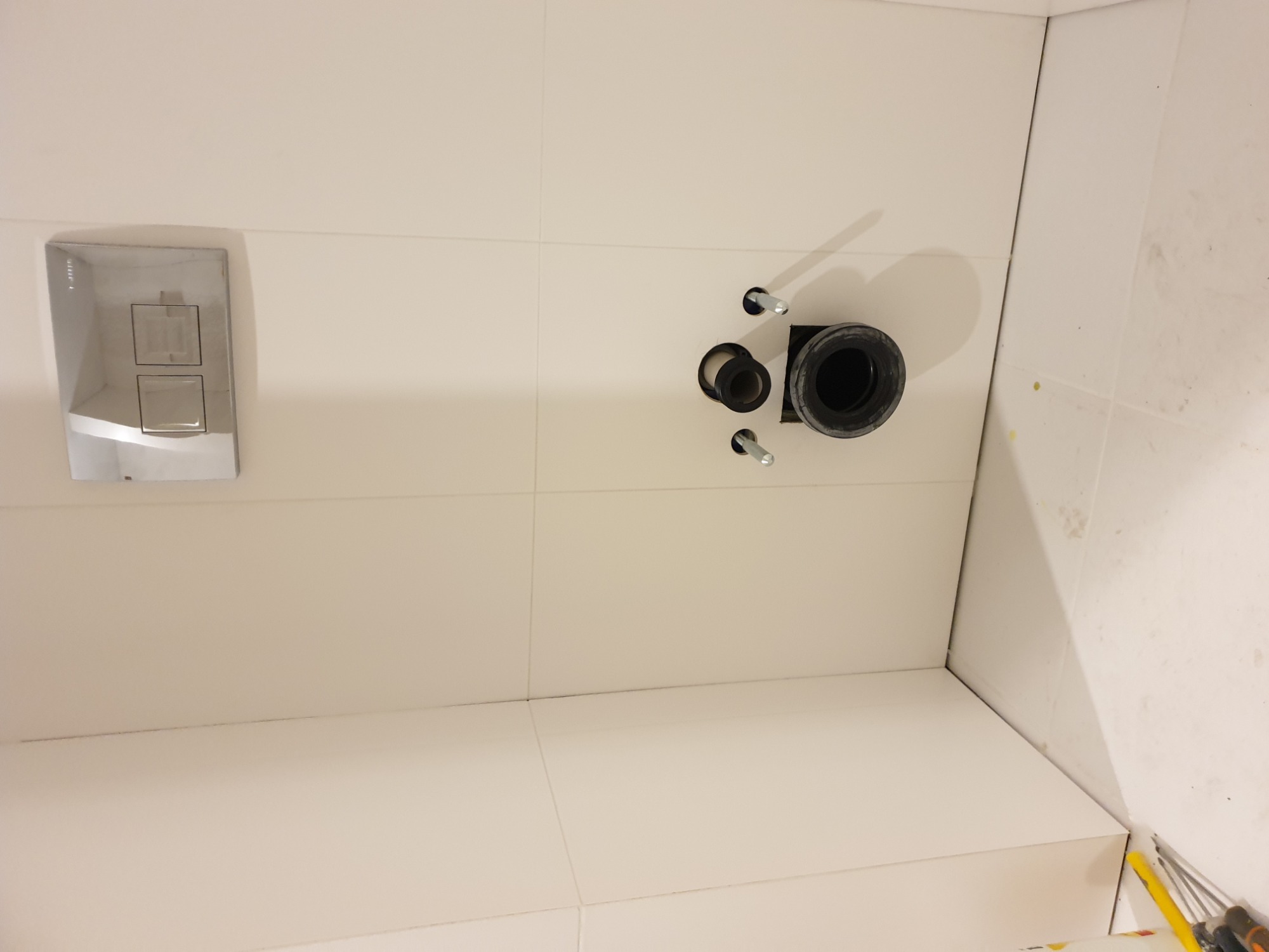 Świeżo wykafelkowana ściana łazienki z zamontowanym stelażem podtynkowym WC, widoczne wystające śruby mocujące i otwór kanalizacyjny.