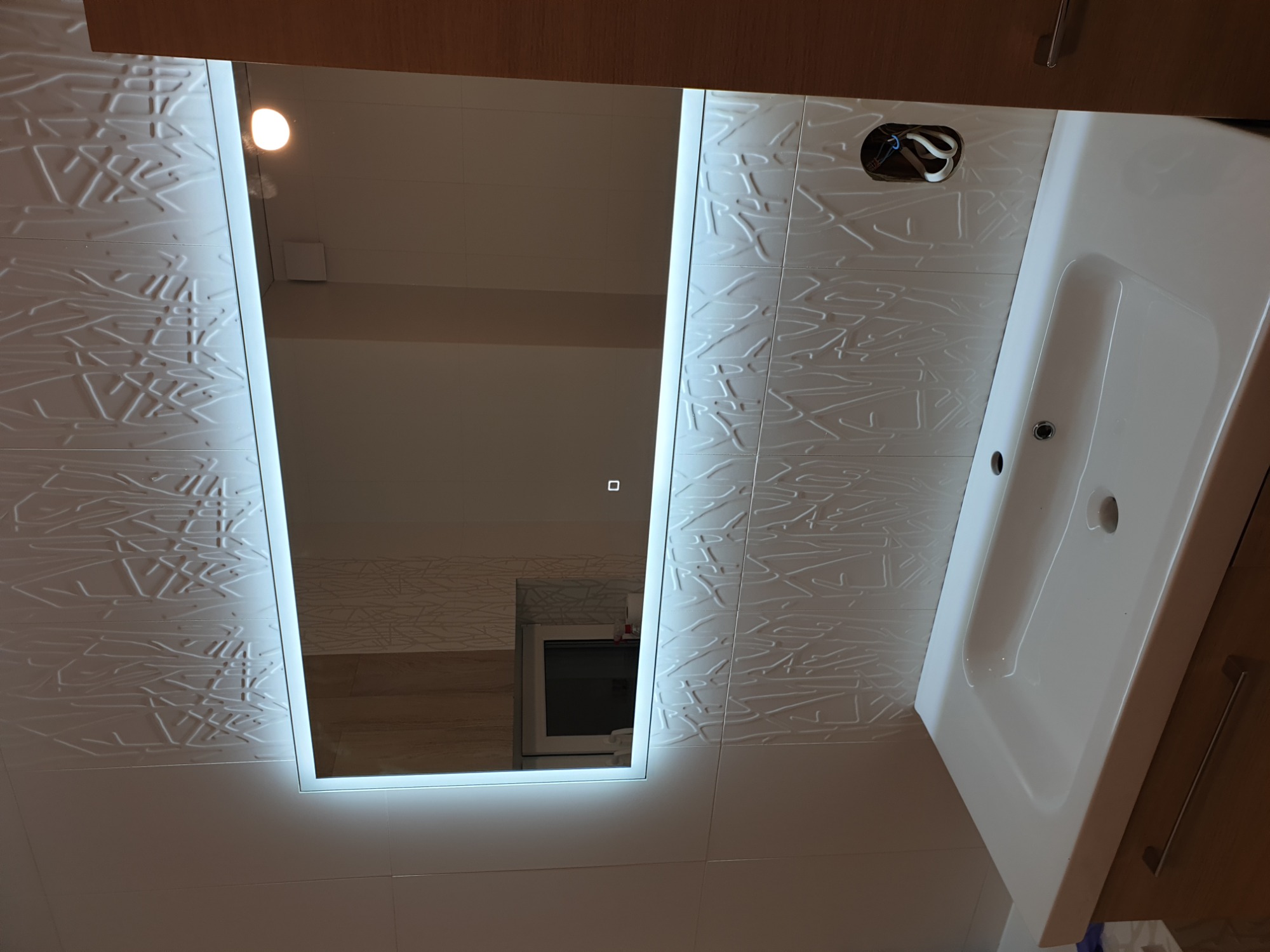 Nowoczesna łazienka z podświetlanym lustrem i umywalką, płytki ścienne z delikatnym wzorem, widoczne niedokończone podłączenie elektryczne.