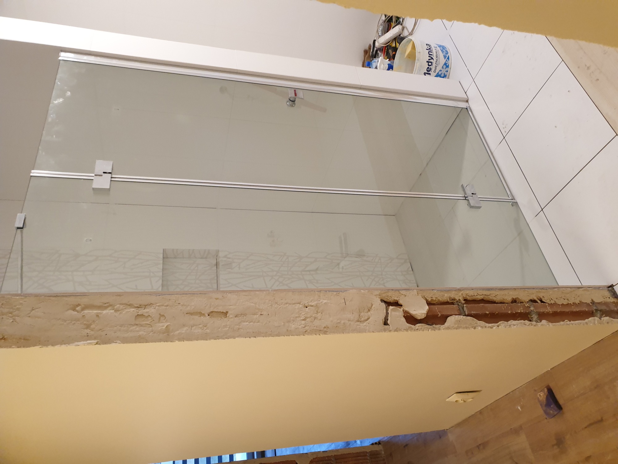 Szklana kabina prysznicowa z chromowanymi okuciami, zamontowana w łazience w trakcie remontu, widoczne niedokończone wykończenia ścian i sufitu oraz białe płytki na ścianie.