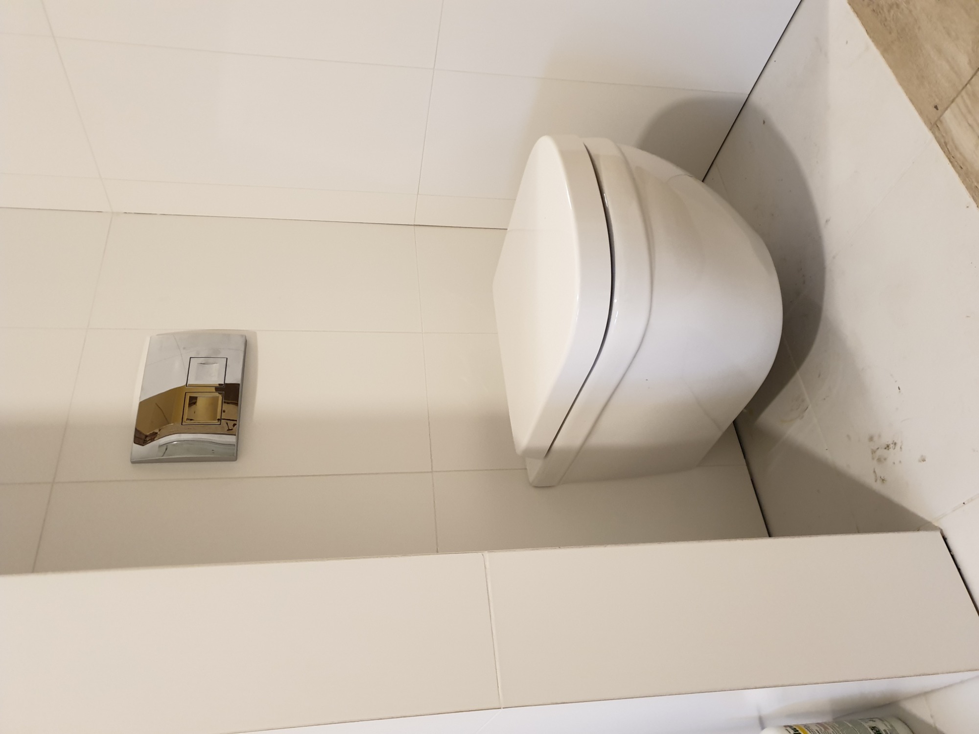 Nowoczesna, podwieszana toaleta w jasnej łazience z białymi płytkami, widoczny chromowany przycisk spłukujący i fragment podłogi z jasnymi płytkami imitującymi drewno.