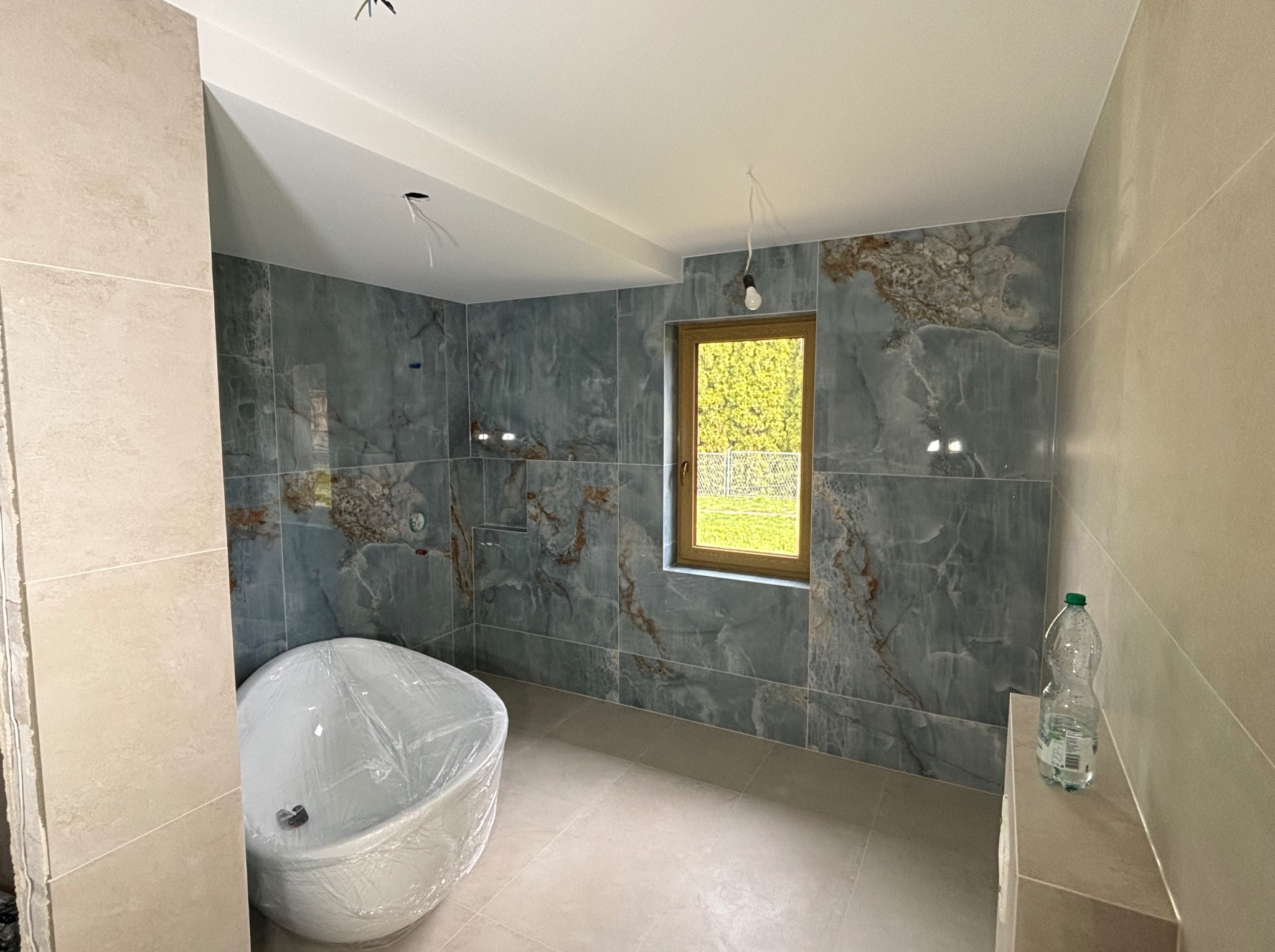 Niedokończona łazienka z niebiesko-szarymi płytkami imitującymi marmur, zabezpieczona folią wanna i butelka wody na półce.