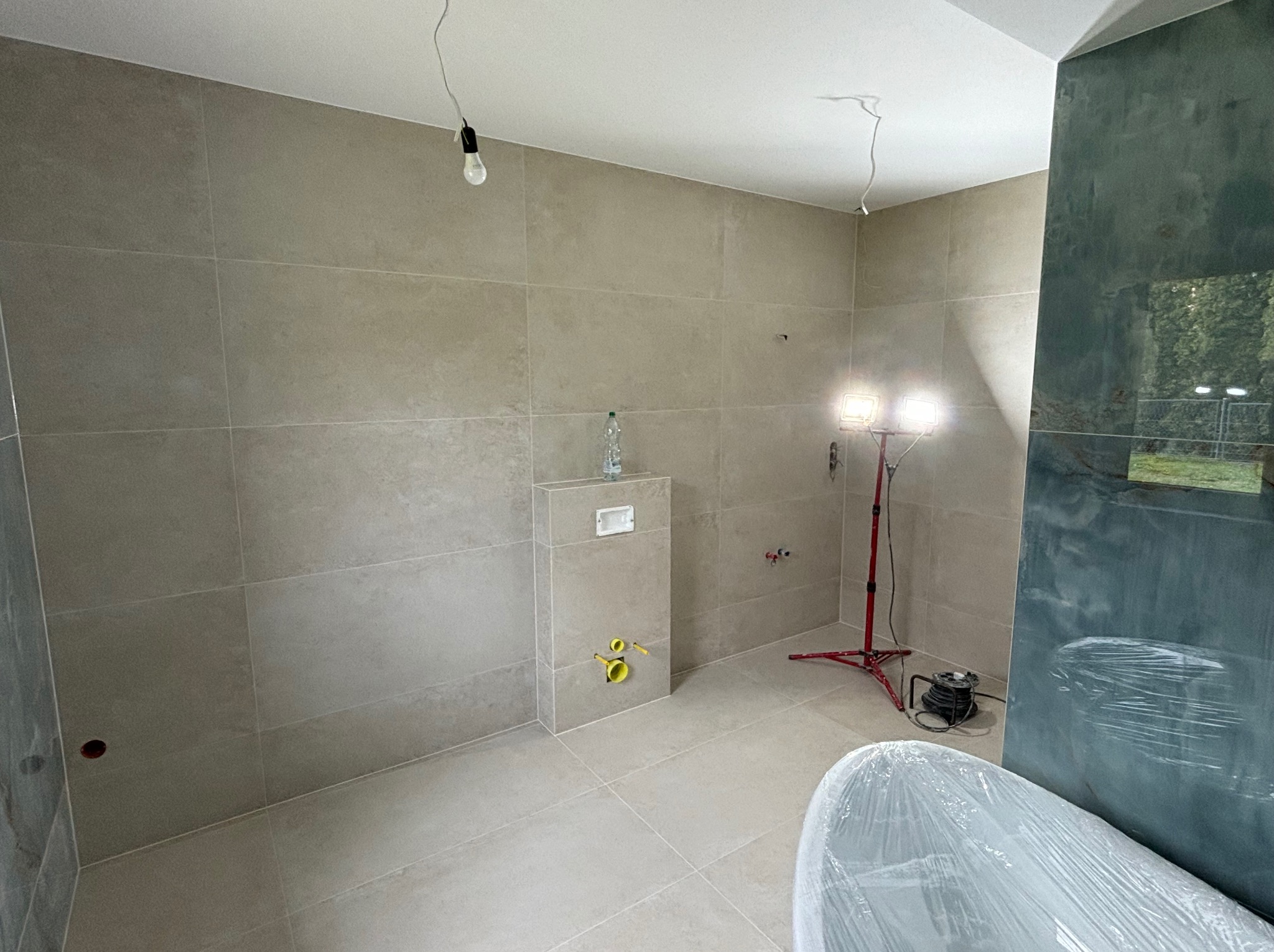 Wnętrze łazienki w trakcie prac: beżowe płytki na ścianach i podłodze, zabudowa podtynkowego WC, tymczasowe oświetlenie halogenowe na statywie, nowa wanna zabezpieczona folią, częściowo przeszklona...