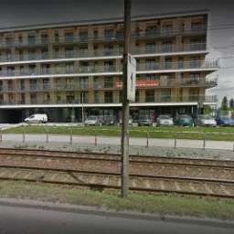 Wielopiętrowy budynek mieszkalny z balkonami, widoczny z poziomu torów kolejowych, z zaparkowanymi samochodami i reklamą na fasadzie.