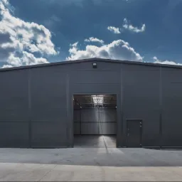Nowoczesny, szary budynek magazynowy z otwartą bramą wjazdową i małym wejściem bocznym, betonowy plac przed budynkiem, błękitne niebo z chmurami w tle.