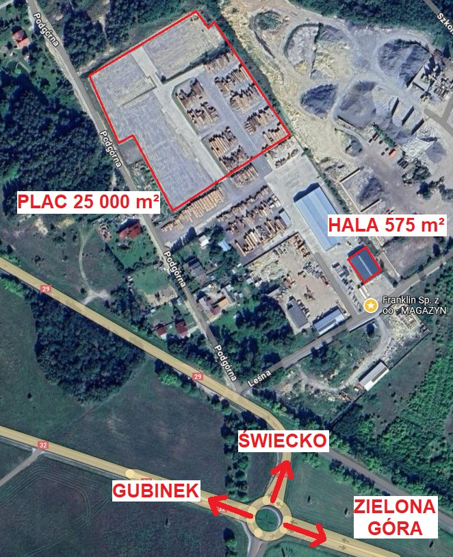 Zdjęcie z lotu ptaka przedstawiające plac składowy z ułożonym drewnem, przylegającą halą i drogą dojazdową z rondem.